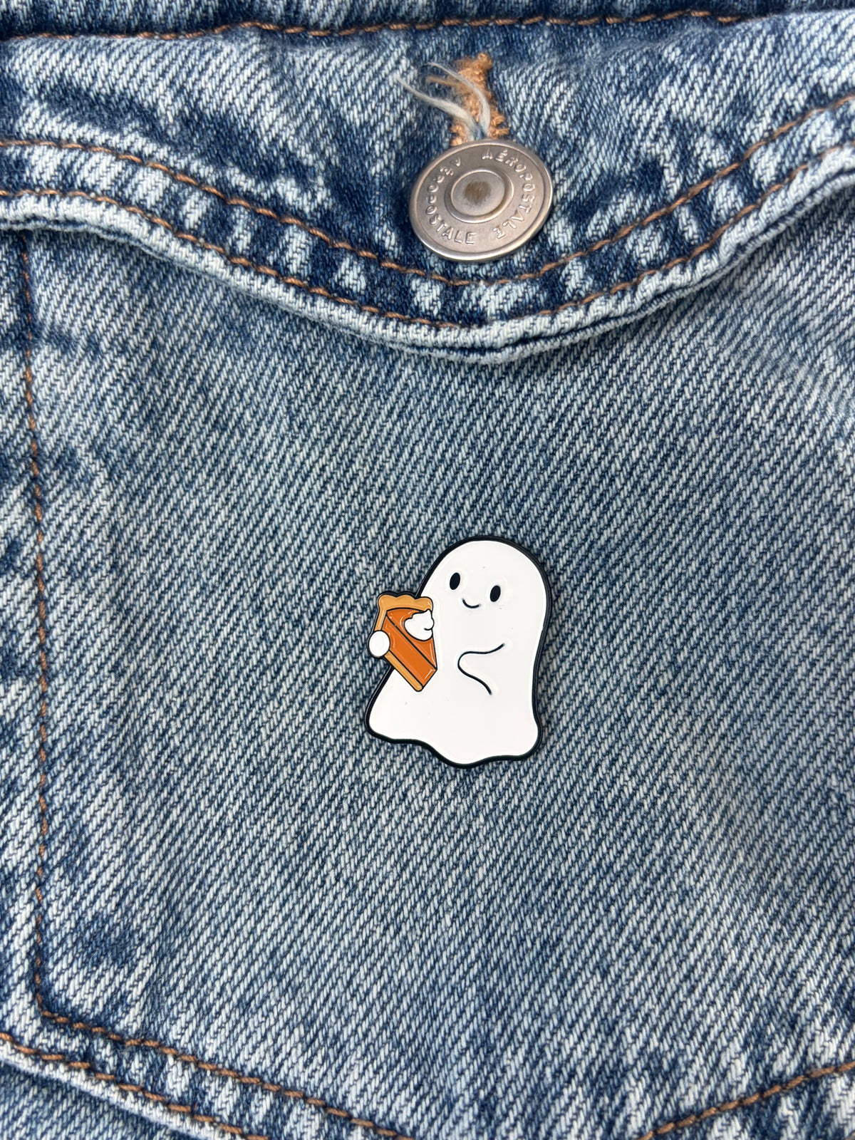 A small enamel pin shaped like a white cartoon ghost holding a pumpkin pie slice, placed on blue denim jeans.